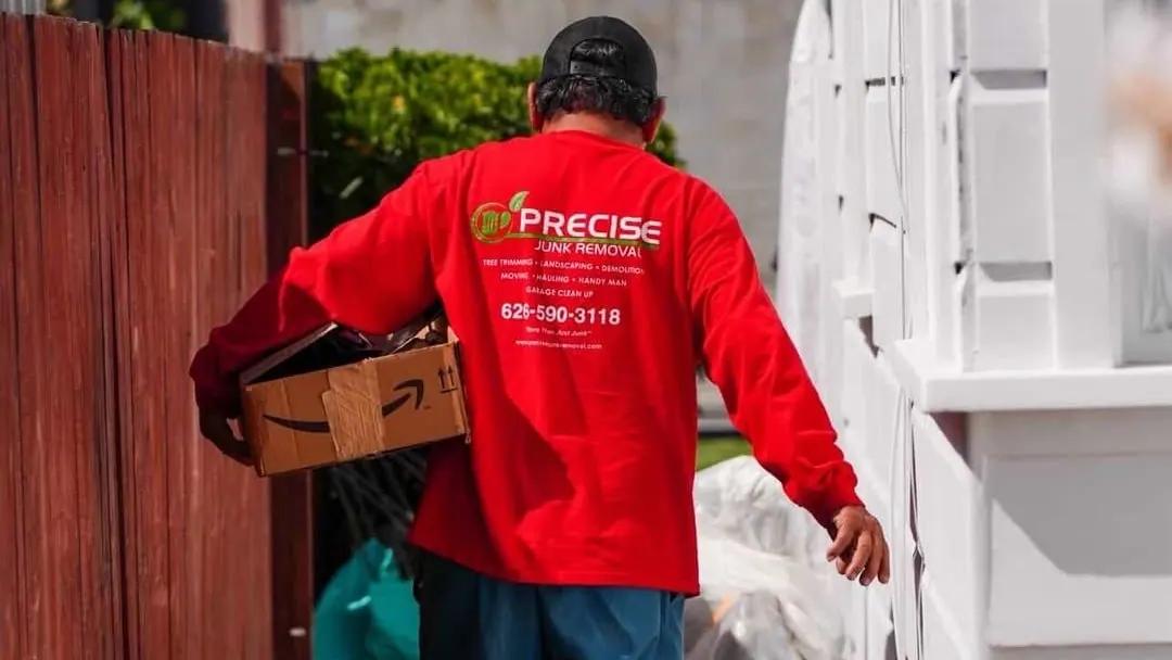 A crew member in a red Precise shirt carrying a cardboard box along a side path