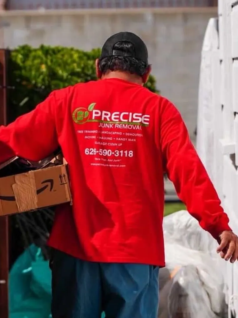 A crew member in a red Precise shirt carrying a cardboard box along a side path