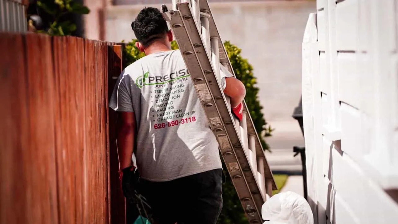 A crew member carrying an aluminum extension ladder along a path beside a house