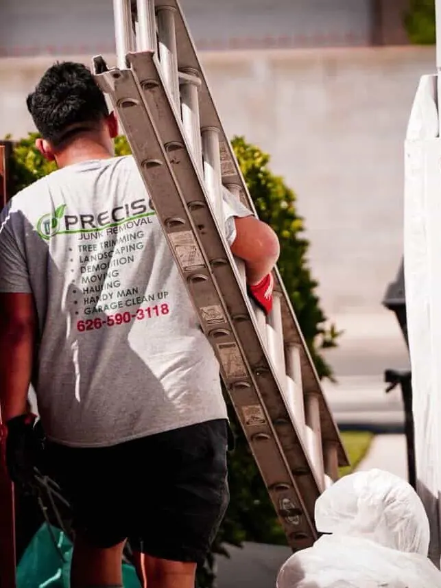 A crew member carrying an aluminum extension ladder along a path beside a house