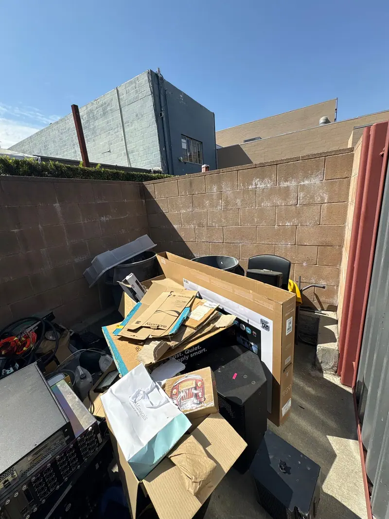 A walled corner stacked with a flat screen TV box, speakers, cords and an old rack of equipment