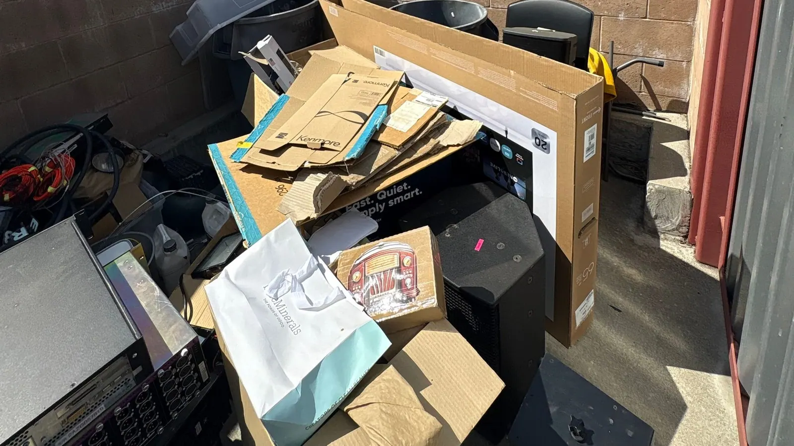 A pile of electronics waiting for pickup: a flat screen TV box, speakers, cords and an old rack of equipment