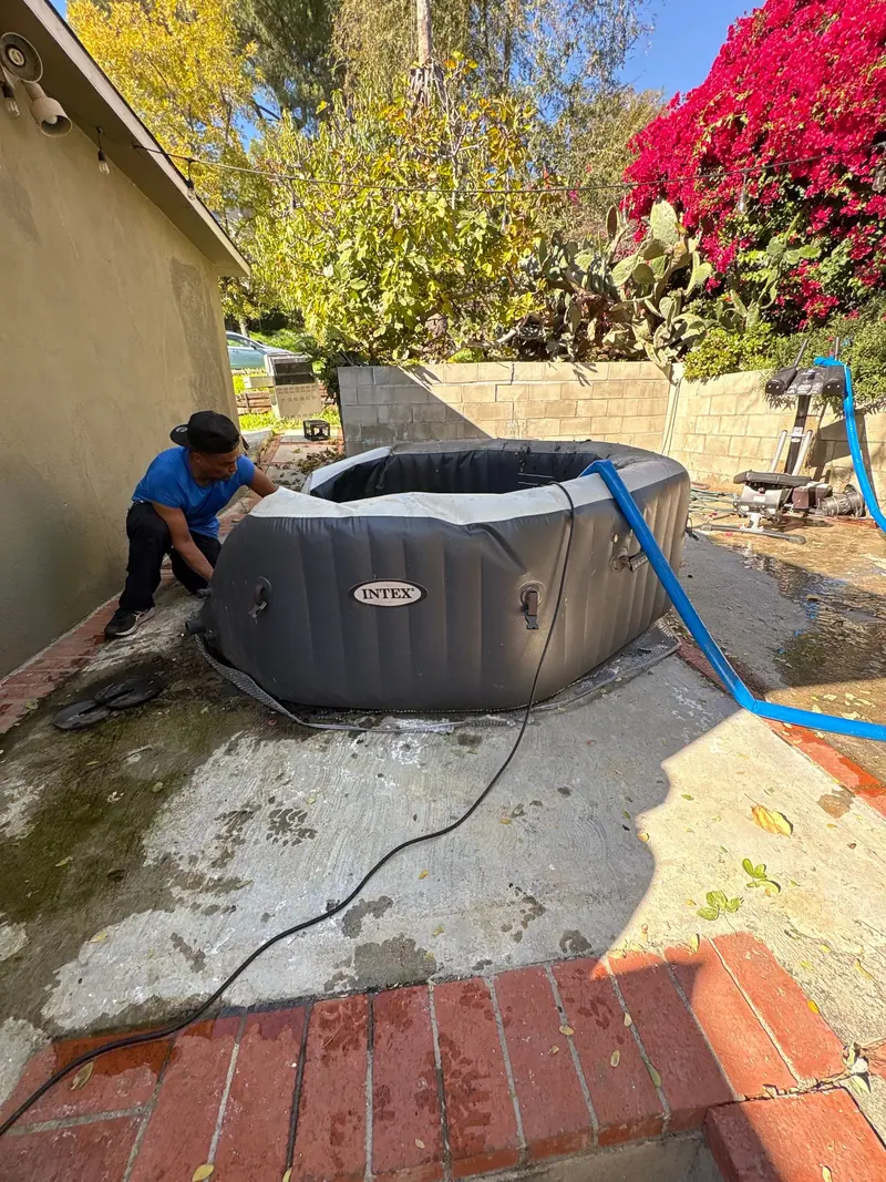 A crew member folding down an inflatable hot tub after draining it, the drain hose still running