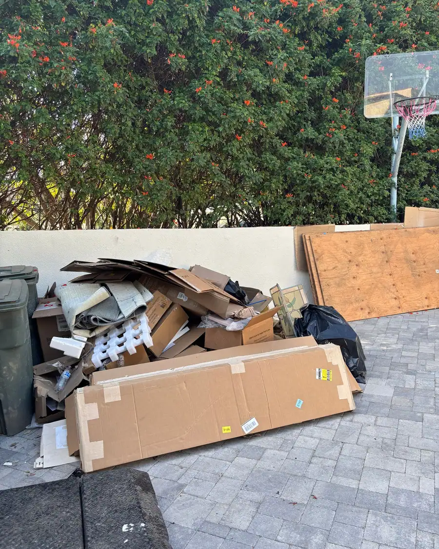 Flattened cartons, a rolled carpet offcut, foam packing and plywood sheets piled against a wall on a paver driveway, beside the wheeled bins